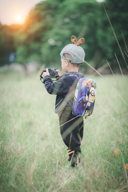 Boy Carrying Backpack Holding Camera Standing on Green Grass