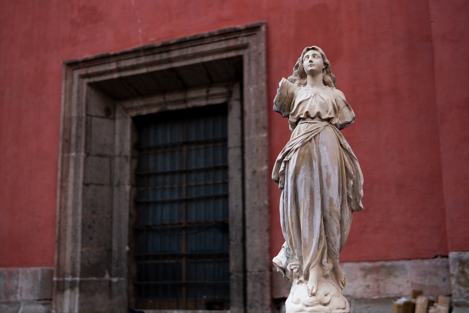 A marble statue stands before a red wall in urban Mexico City, illustrating religious art.