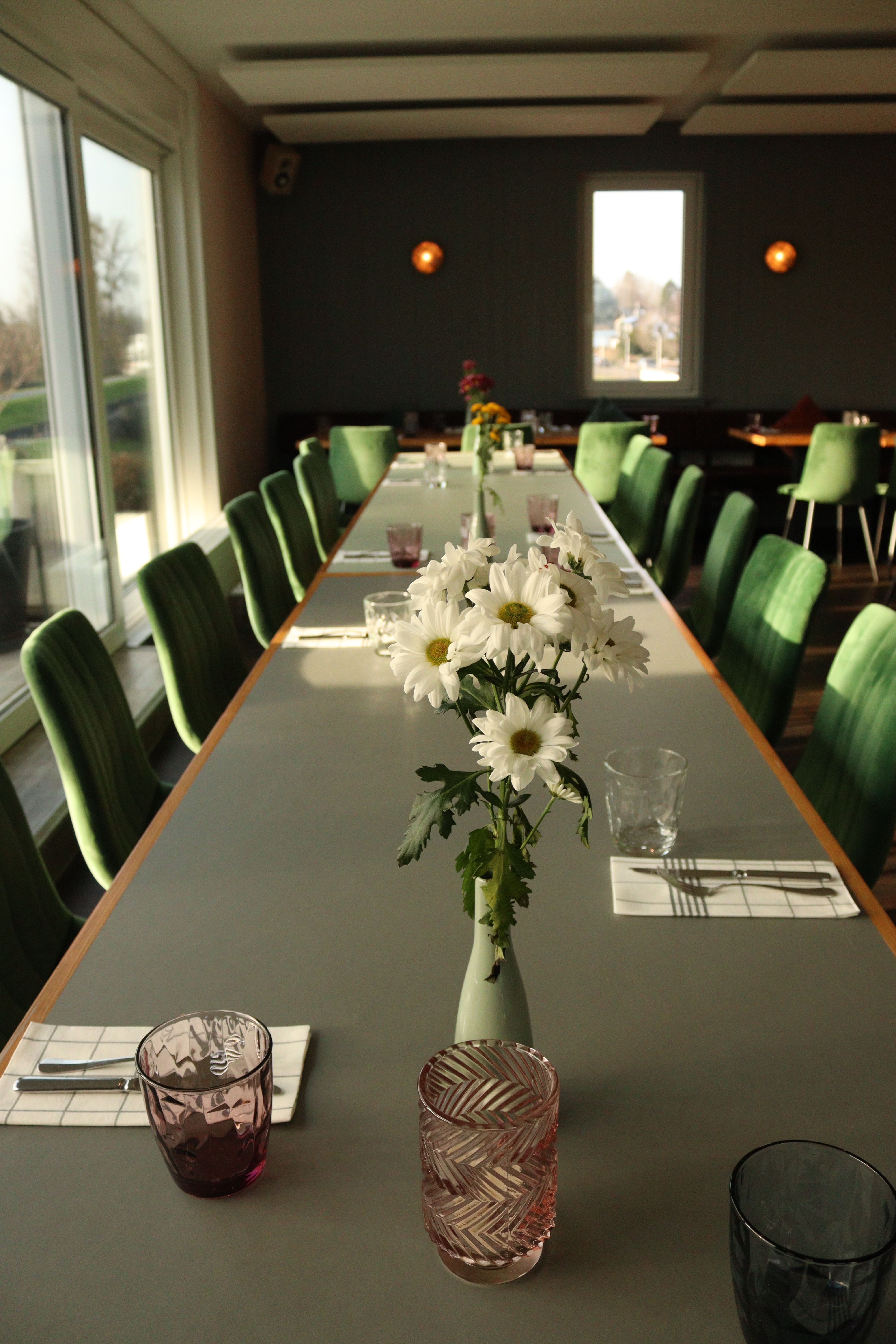 Long dining table with elegant floral centerpiece in a sunlit restaurant with green chairs.