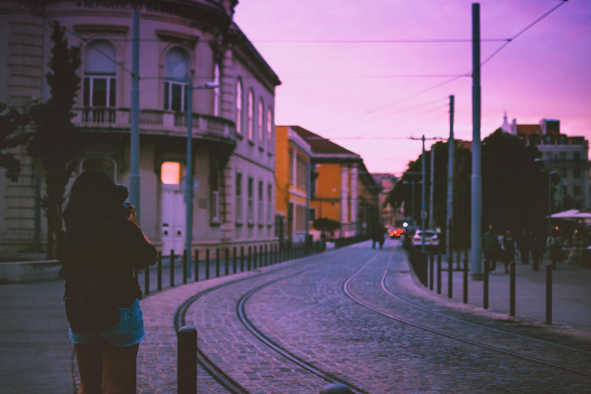 A woman photographs a scenic street in Lisbon during a vibrant sunset with tram lines.