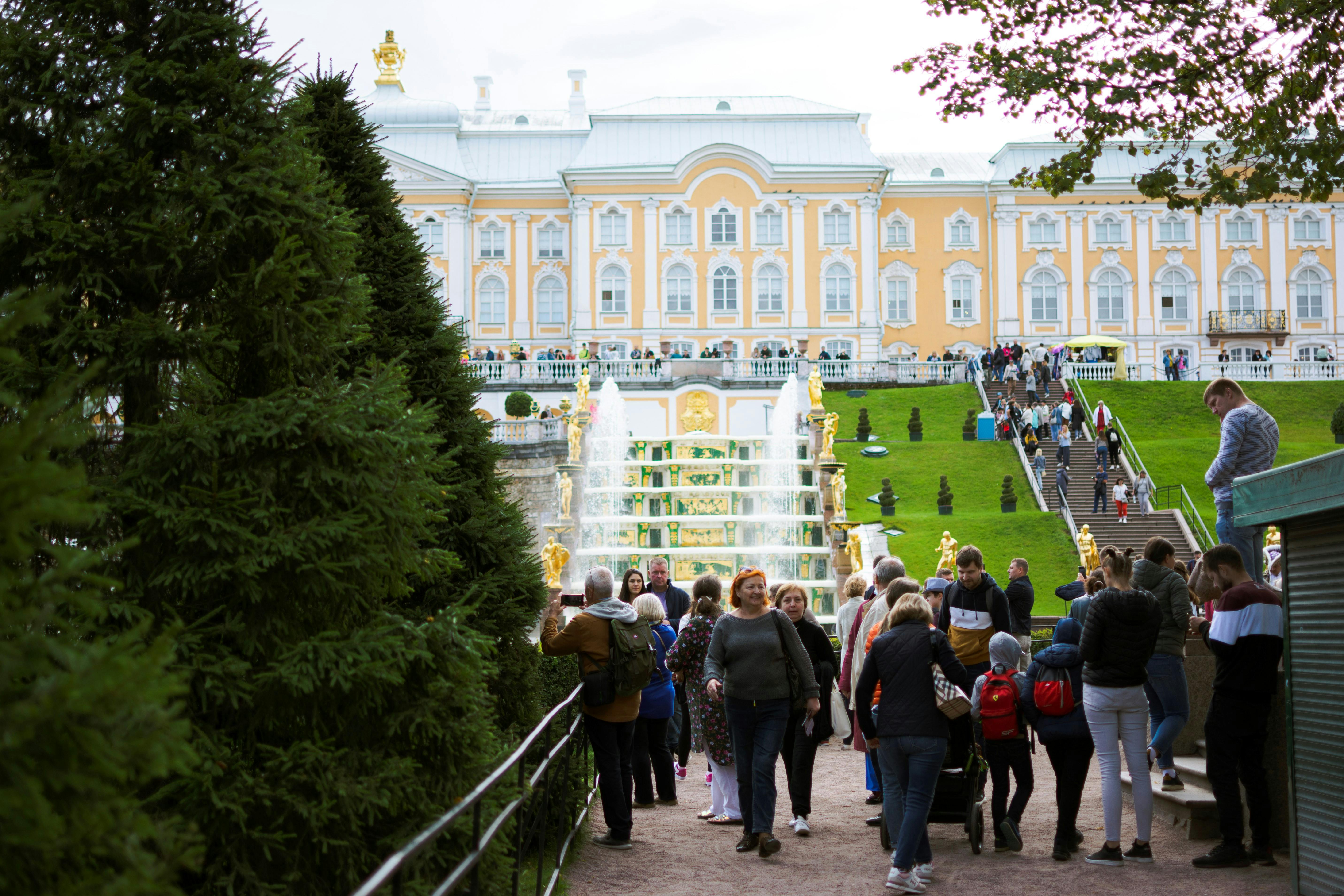 People gather at the Grand Cascade fountain in front of the Peterhof Palace, enjoying a sunny day in a lush park.