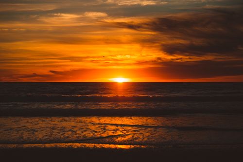 Free A breathtaking sunset over the ocean with vivid orange hues and dramatic clouds reflected in the water. Stock Photo