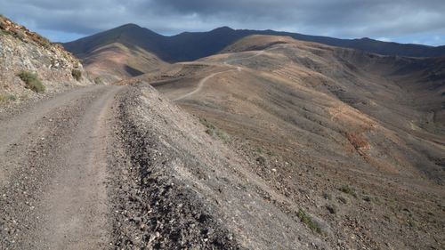 Free Dirt road winds through arid mountain landscape under cloudy sky. Stock Photo