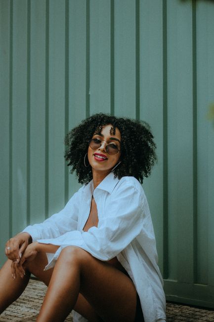 Chic young woman with Afro hairstyle wearing sunglasses and a white shirt, sitting against a green backdrop outdoors.