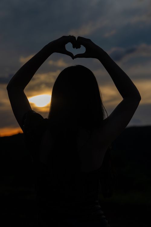 Free Silhouette of a woman forming a heart shape with hands at sunset, symbolizing love and peace. Stock Photo