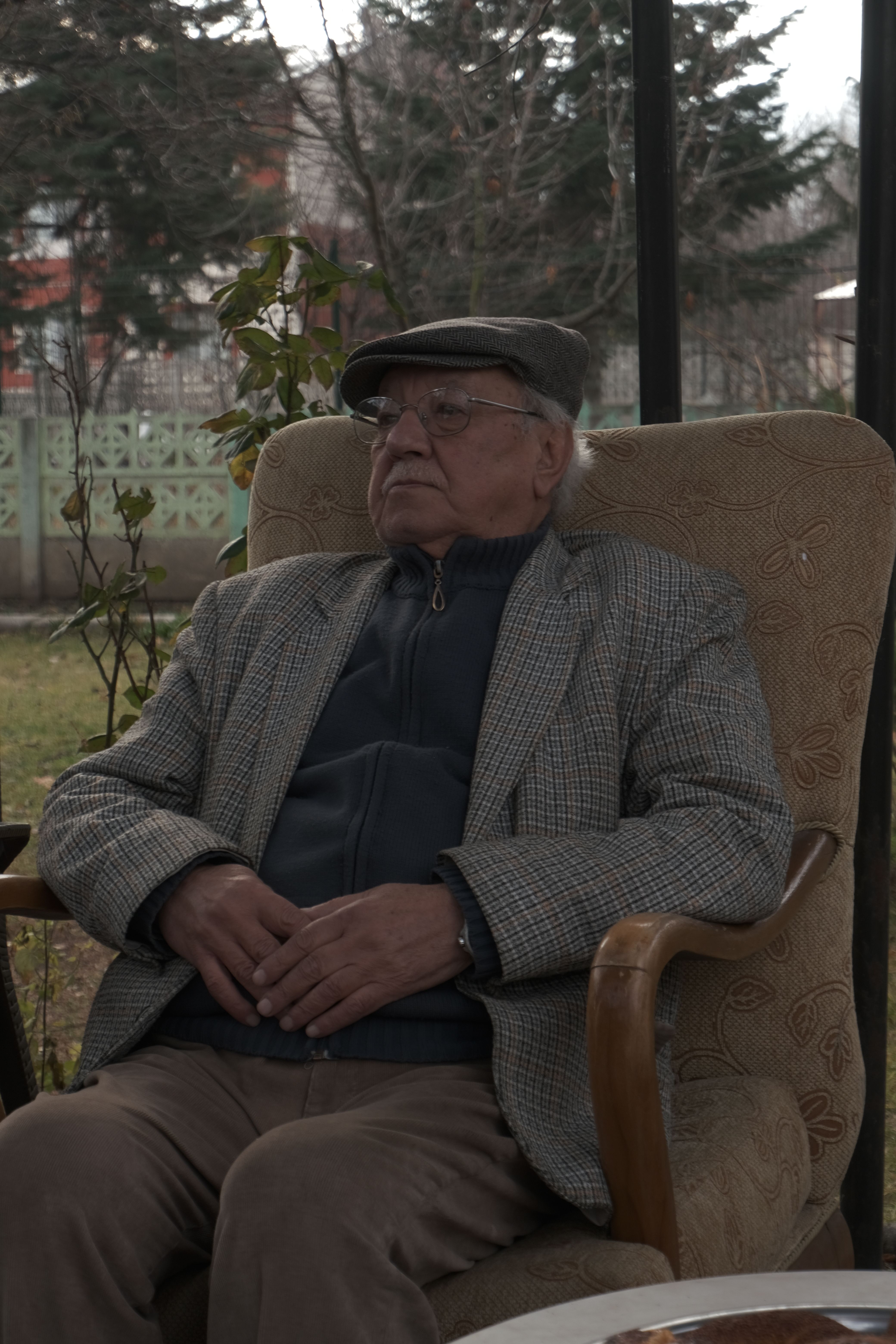 Older man in a flat cap and suit sitting in an armchair outdoors in a garden setting.