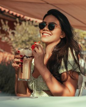 A woman in sunglasses and a green dress enjoys a milkshake outdoors on a sunny day.