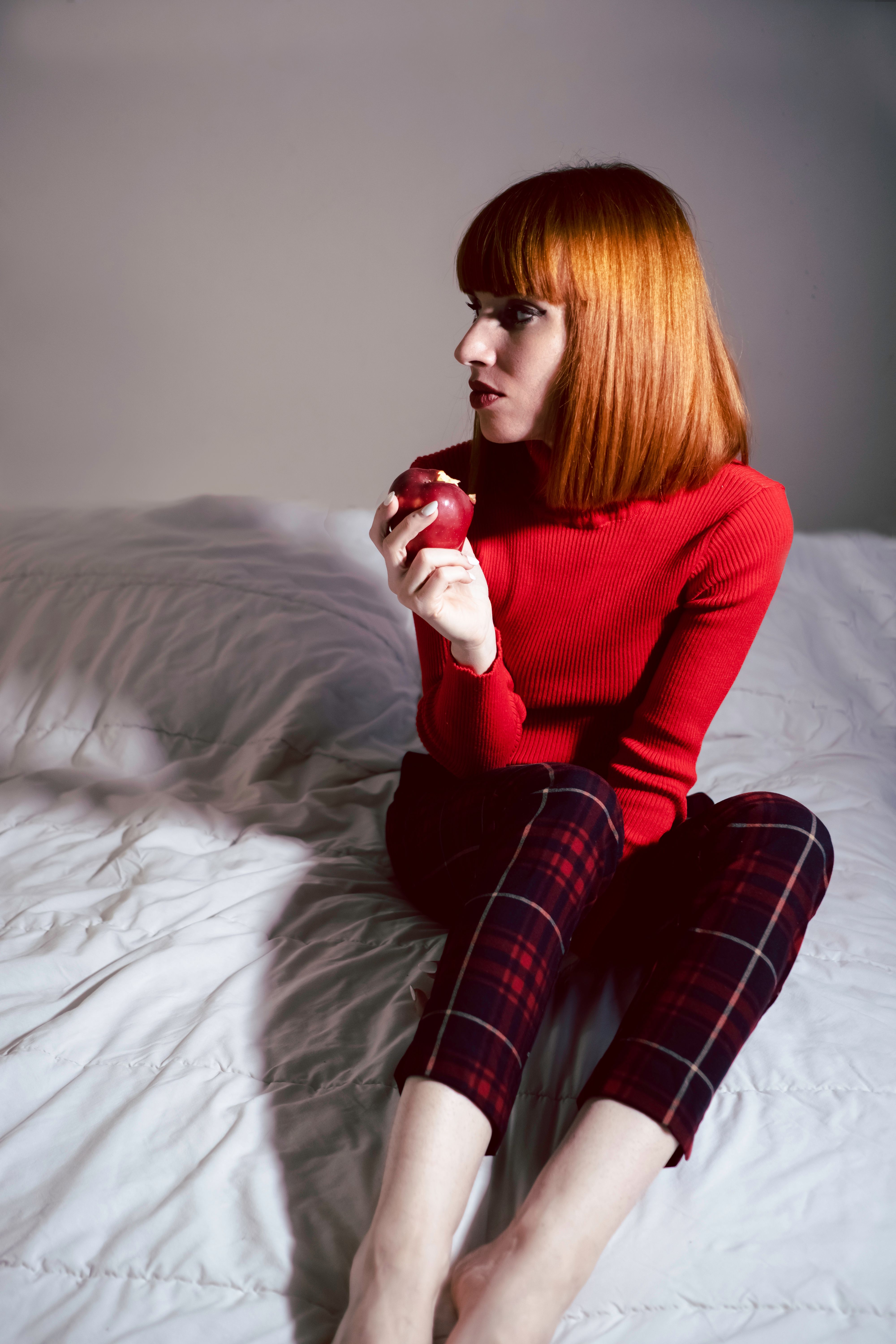 A woman with red hair sitting on a bed holding an apple · Free Stock Photo