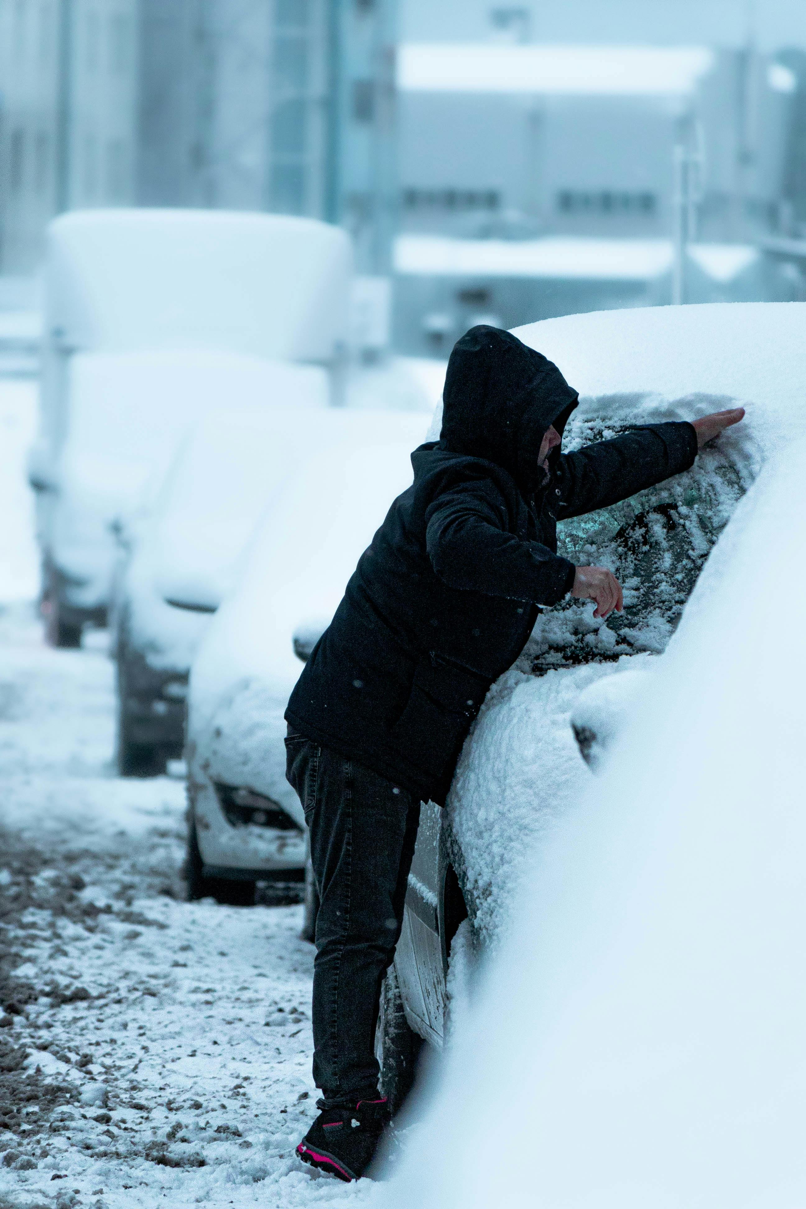 A man clears snow from a car parked on a wintery street, highlighting seasonal car maintenance.