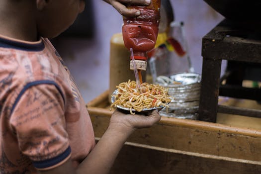 A child pours ketchup over a plate of street food noodles, highlighting vibrant urban flavors.