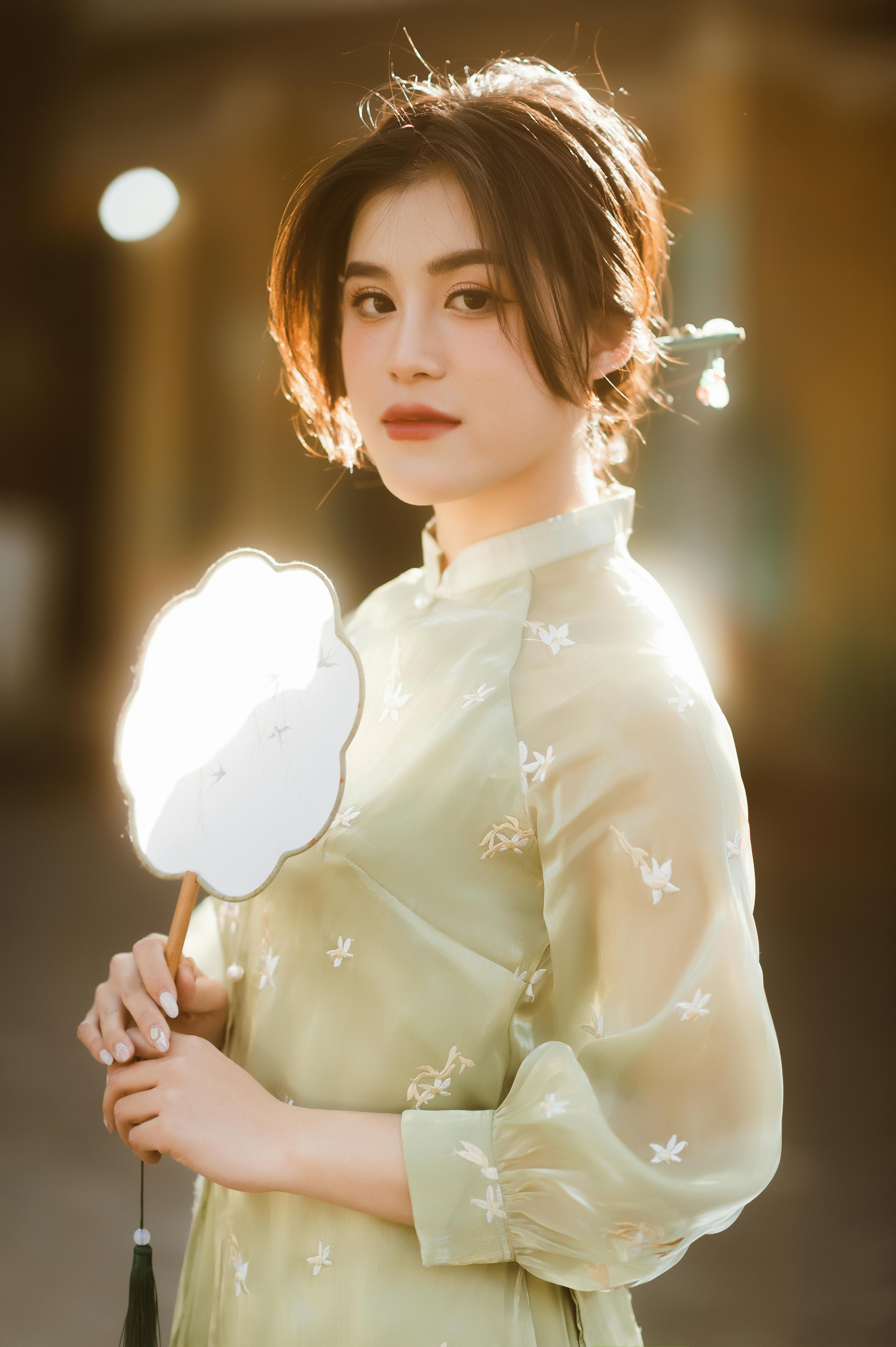 A woman in a traditional dress holding a paper fan, backlit by sunlight, smiling softly.