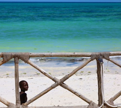 Free A young boy by the ocean in Zanzibar with a rustic wooden fence and clear blue waters. Stock Photo