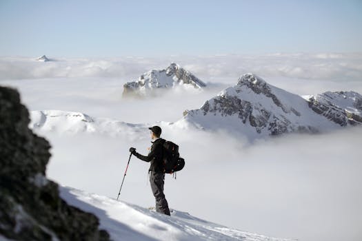 A climber stands confidently overlooking a snowy mountain landscape with clouds.