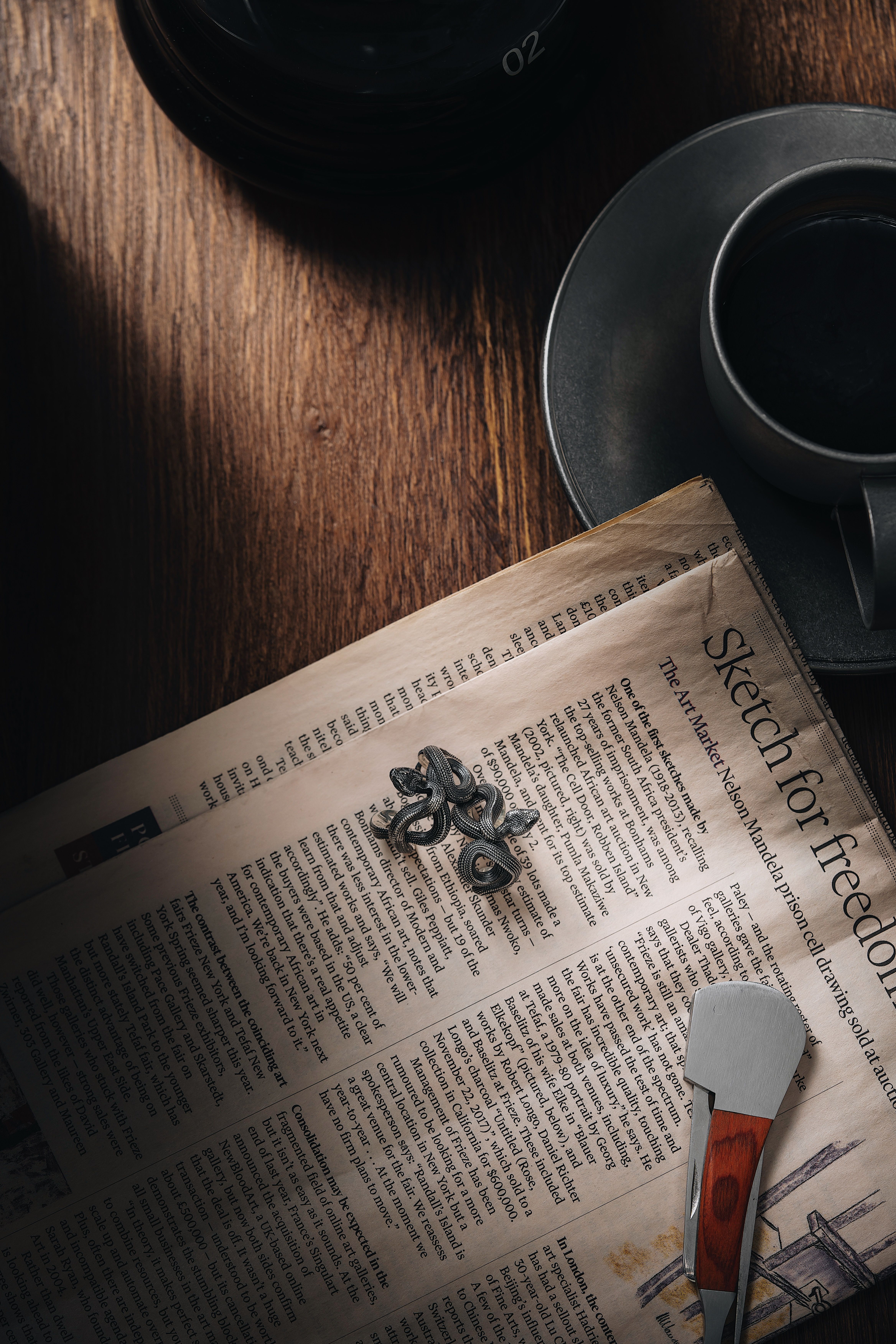 A vintage still life scene featuring a coffee cup, newspaper, and metal ring on a wooden table.
