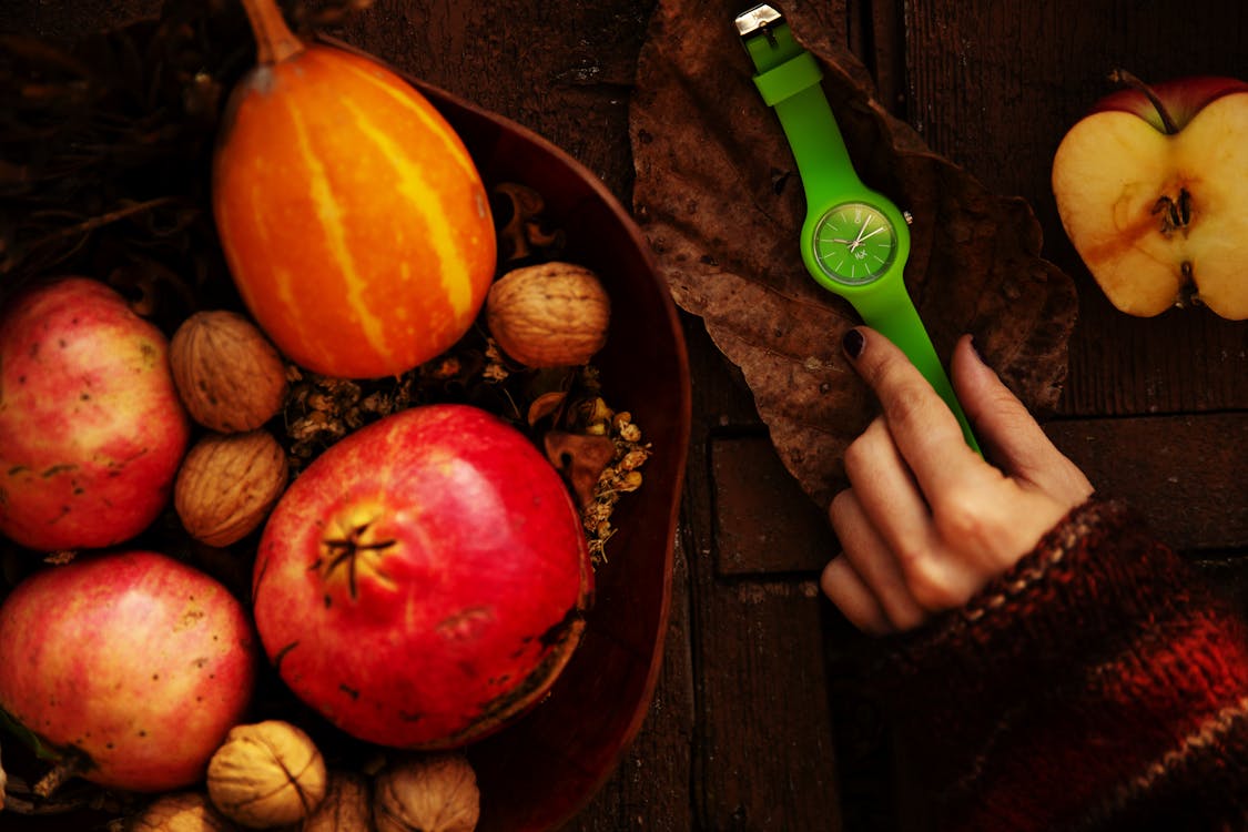 Top view of crop unrecognizable female putting green wristwatch on dry leaf while arranging creative composition with ripe fresh fruits and walnuts