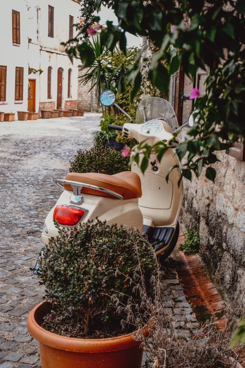 Free Vintage scooter parked in a cozy cobblestone alley surrounded by rustic buildings and lush plants. Stock Photo