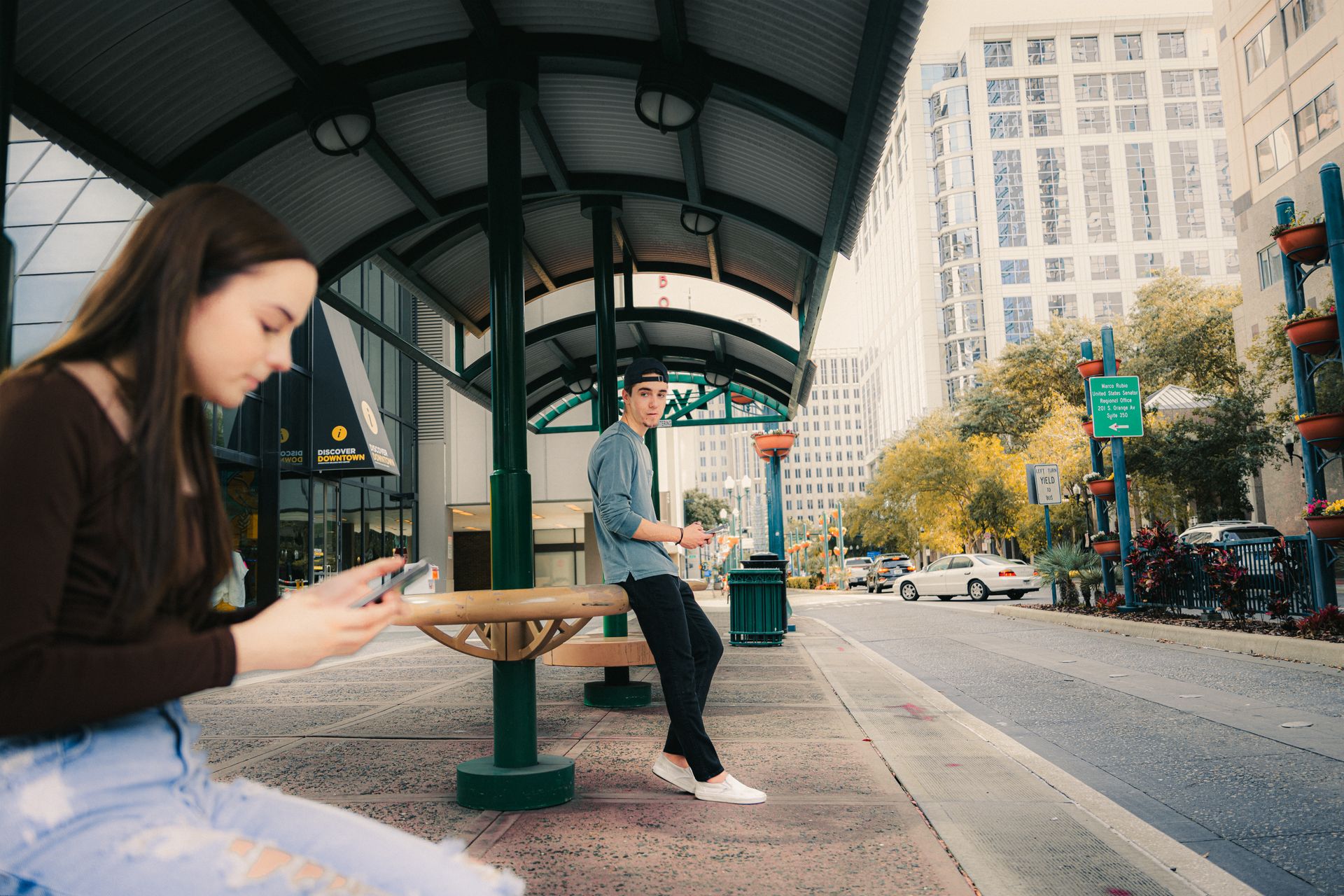 Teens using mobile phones at a city bus stop, urban setting with modern architecture.