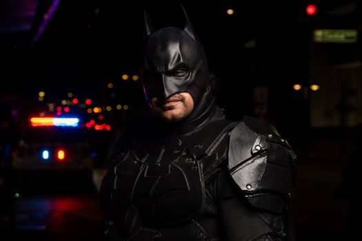 Batman cosplayer in detailed costume at a night event with police lights in the background.