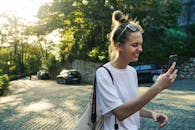 一位快乐的女人在阳光明媚、绿树成荫的鹅卵石街道上查看手机，脸上带着微笑