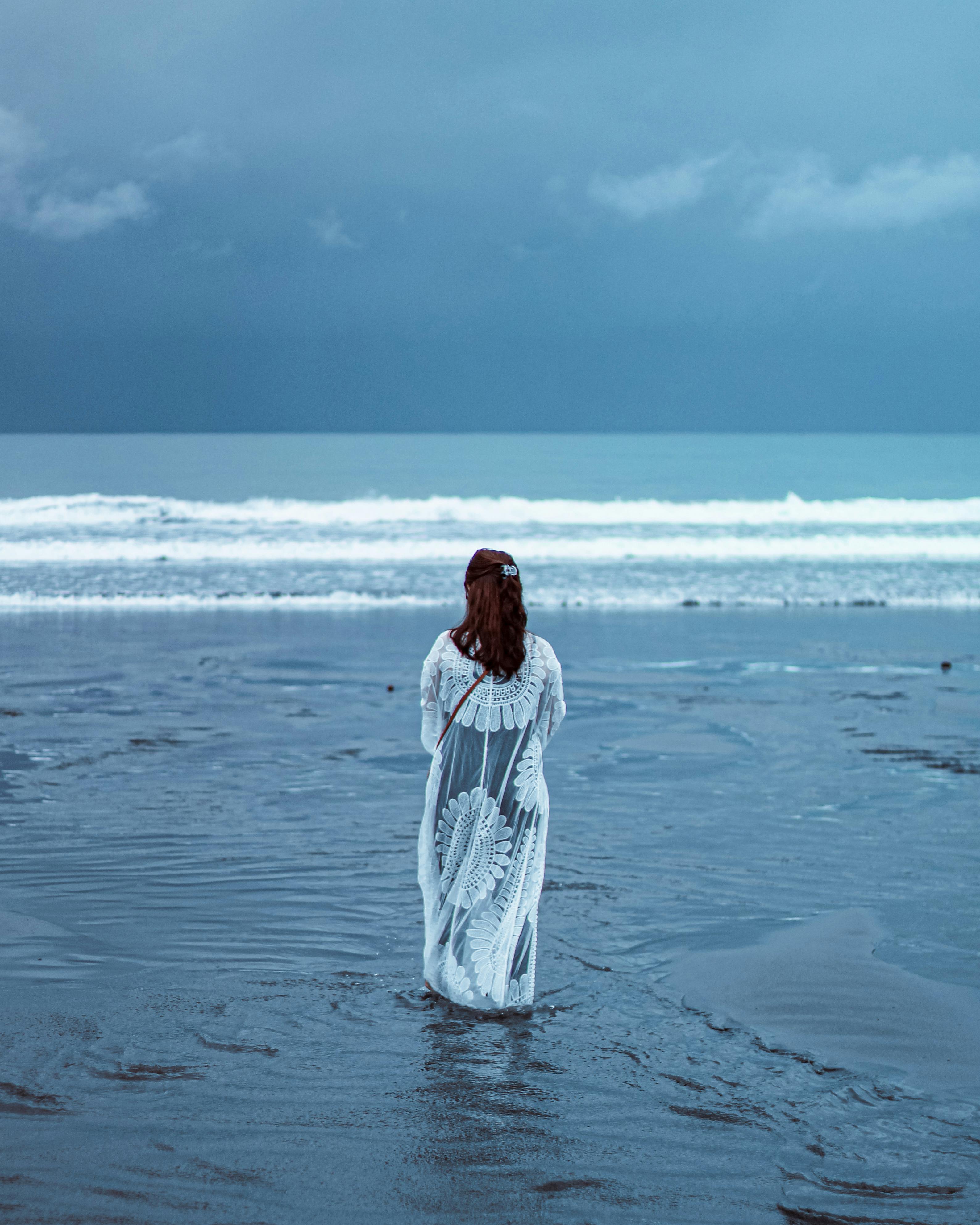 A woman in a white dress stands on a tranquil Indonesian beach, facing the ocean.