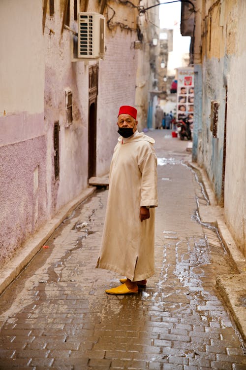 Elderly Man in Gown and Red Hat in Narrow Alley