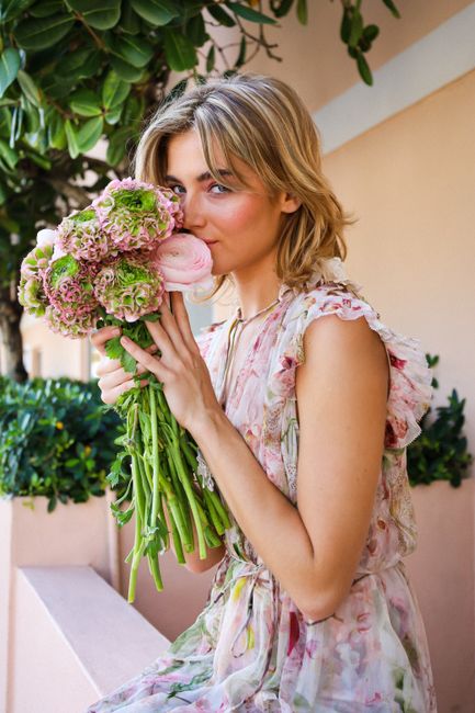 A stylish woman in a floral dress holding a bouquet of flowers near lush greenery.
