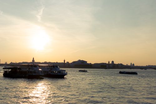 Free Boats sail on tranquil waters as the sun sets over a harbor, creating silhouettes against the evening sky. Stock Photo
