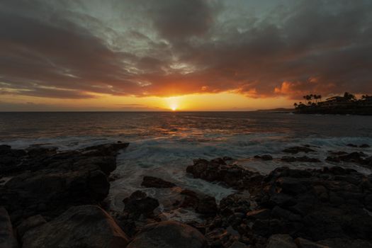 Stunning sunset over a rocky coastline with vibrant colors and silhouetted palms.