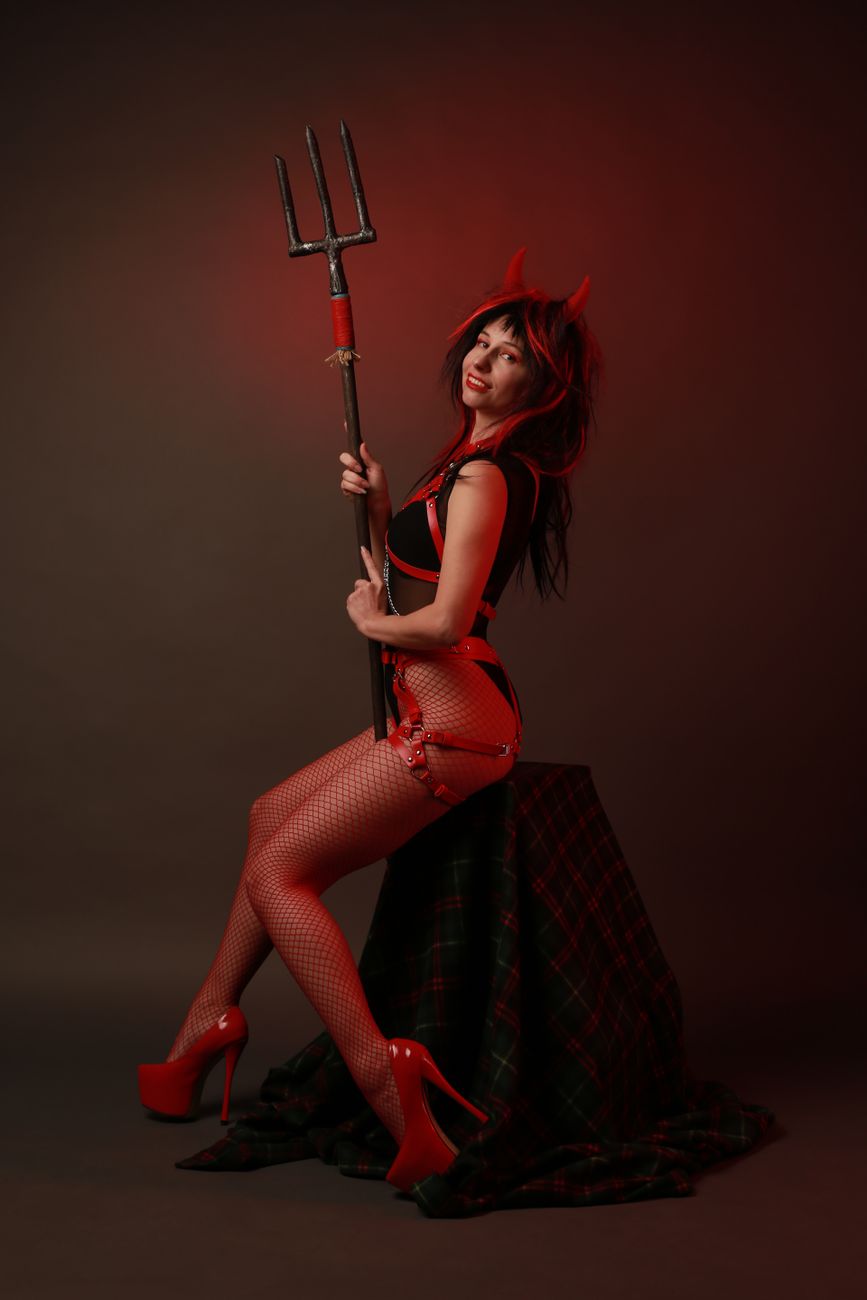 Woman in a devil costume posing with a pitchfork against a red background.