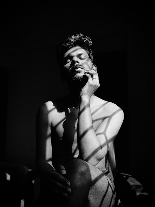 Black and White Photo of a Shirtless Man Sitting in Sunlight 