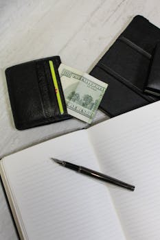 Top-down view of a notebook, pen, wallet, and cash on a light surface, ideal for business or finance themes.