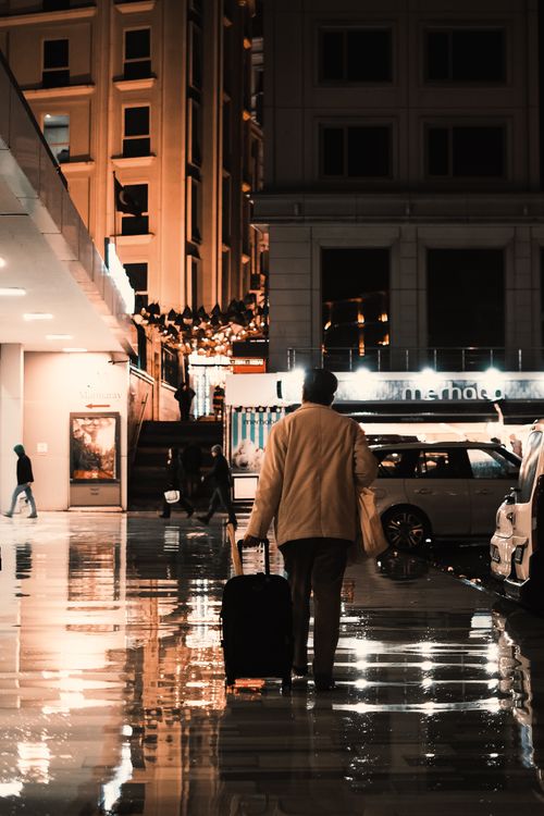 Free A person walking with a suitcase on a wet street at night, reflecting city lights. Stock Photo