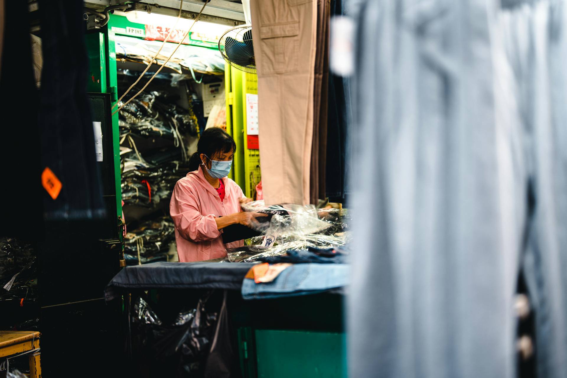 A woman wearing a mask packs clothes in a bustling factory setting.