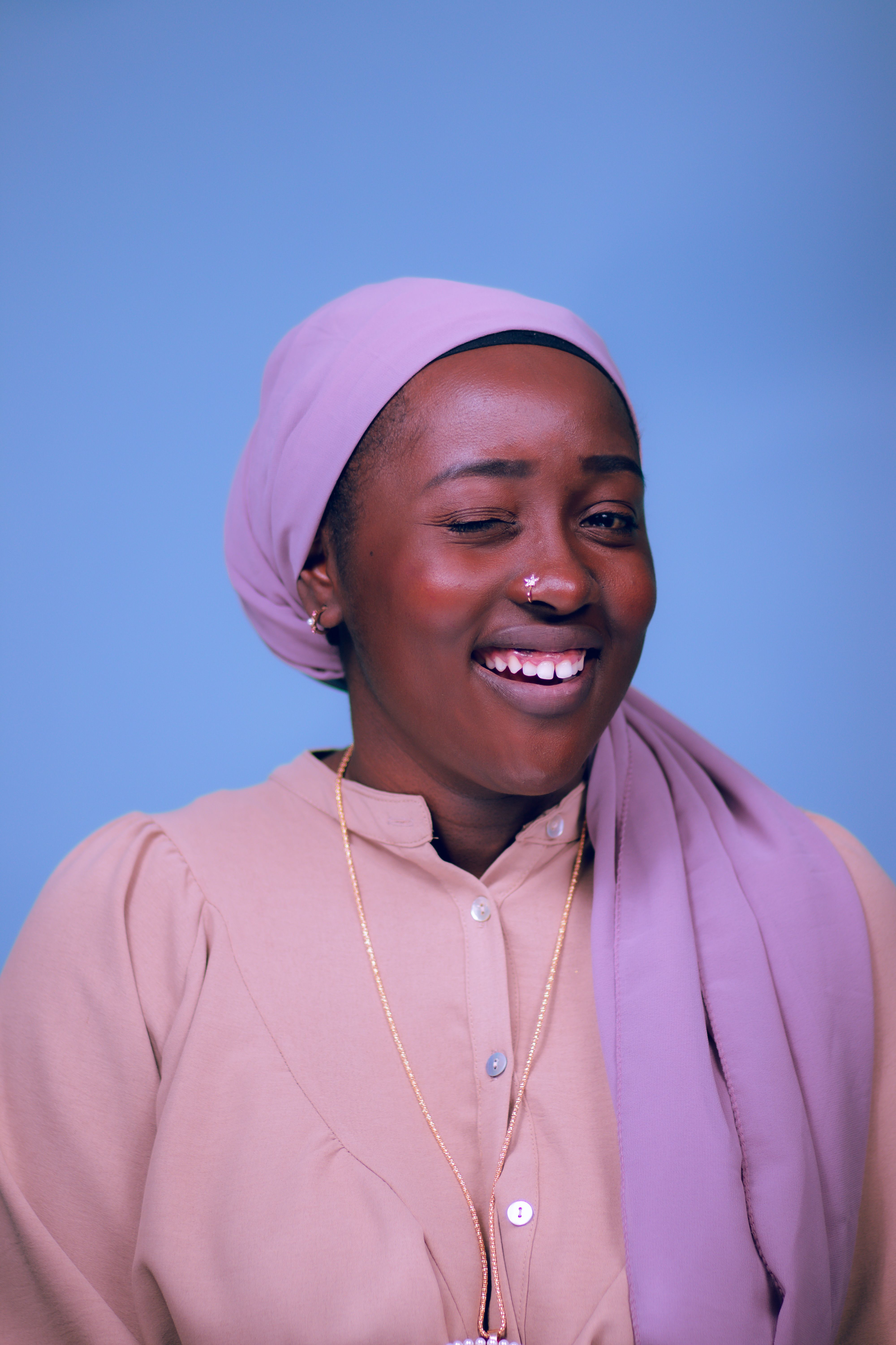 Portrait of a joyful woman wearing a pink headscarf and cream shirt, winking and smiling.
