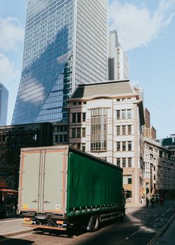 A bustling urban street in London, featuring modern skyscrapers reflecting the sky and a passing truck.