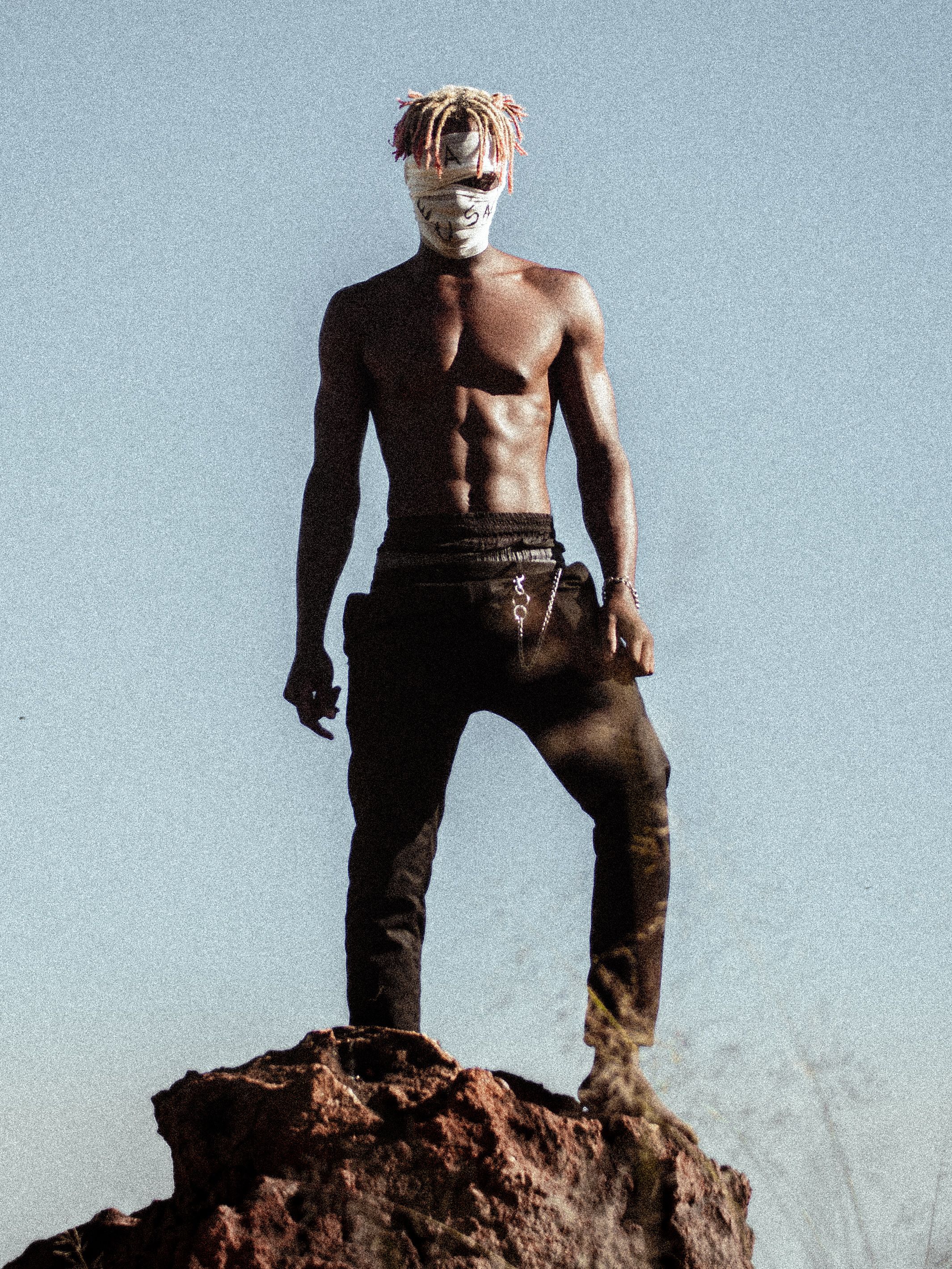 A shirtless man with mask, standing confidently on a rock under a clear sky.