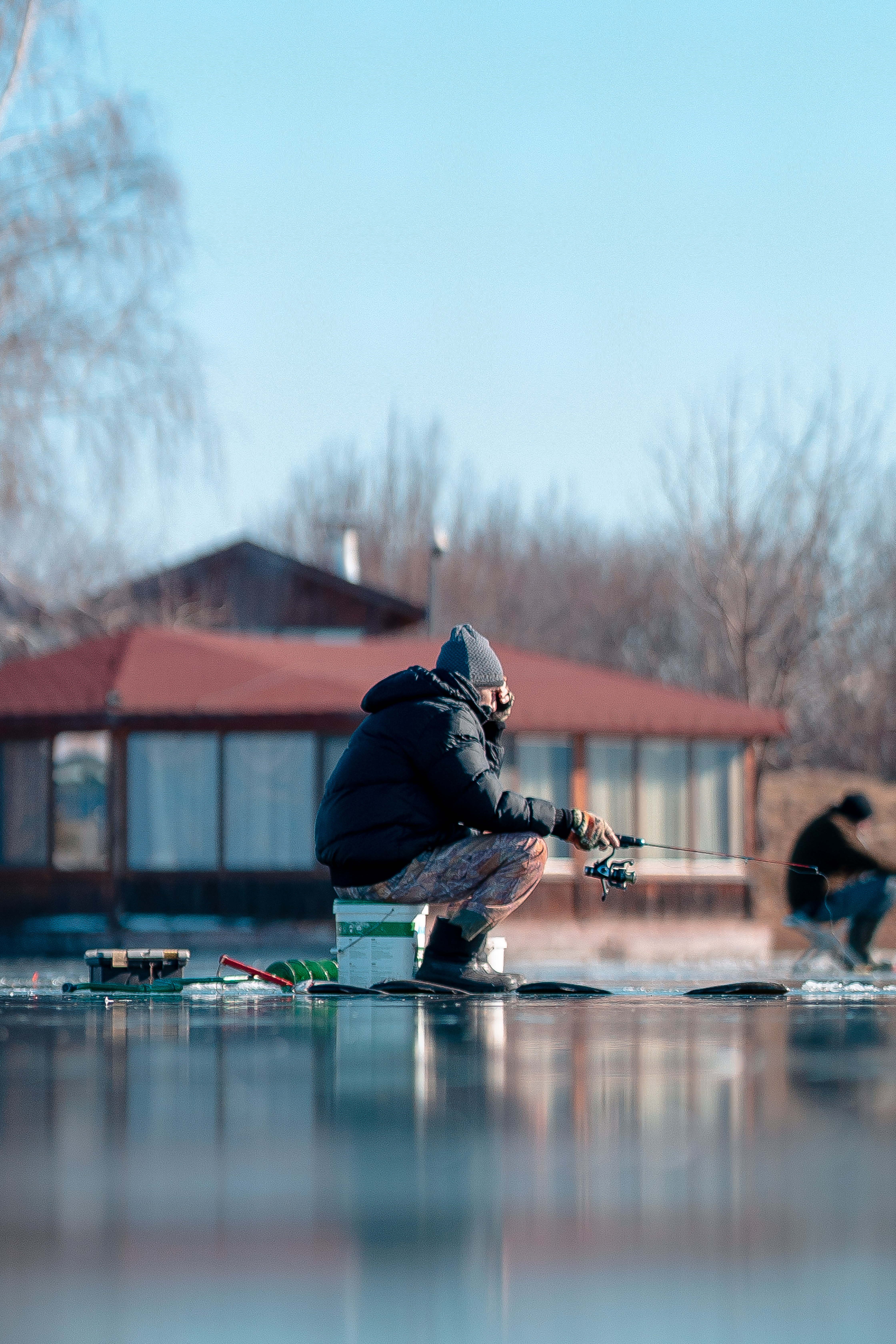 Two people ice fishing on a frozen lake near a cabin. Winter activity outdoors.