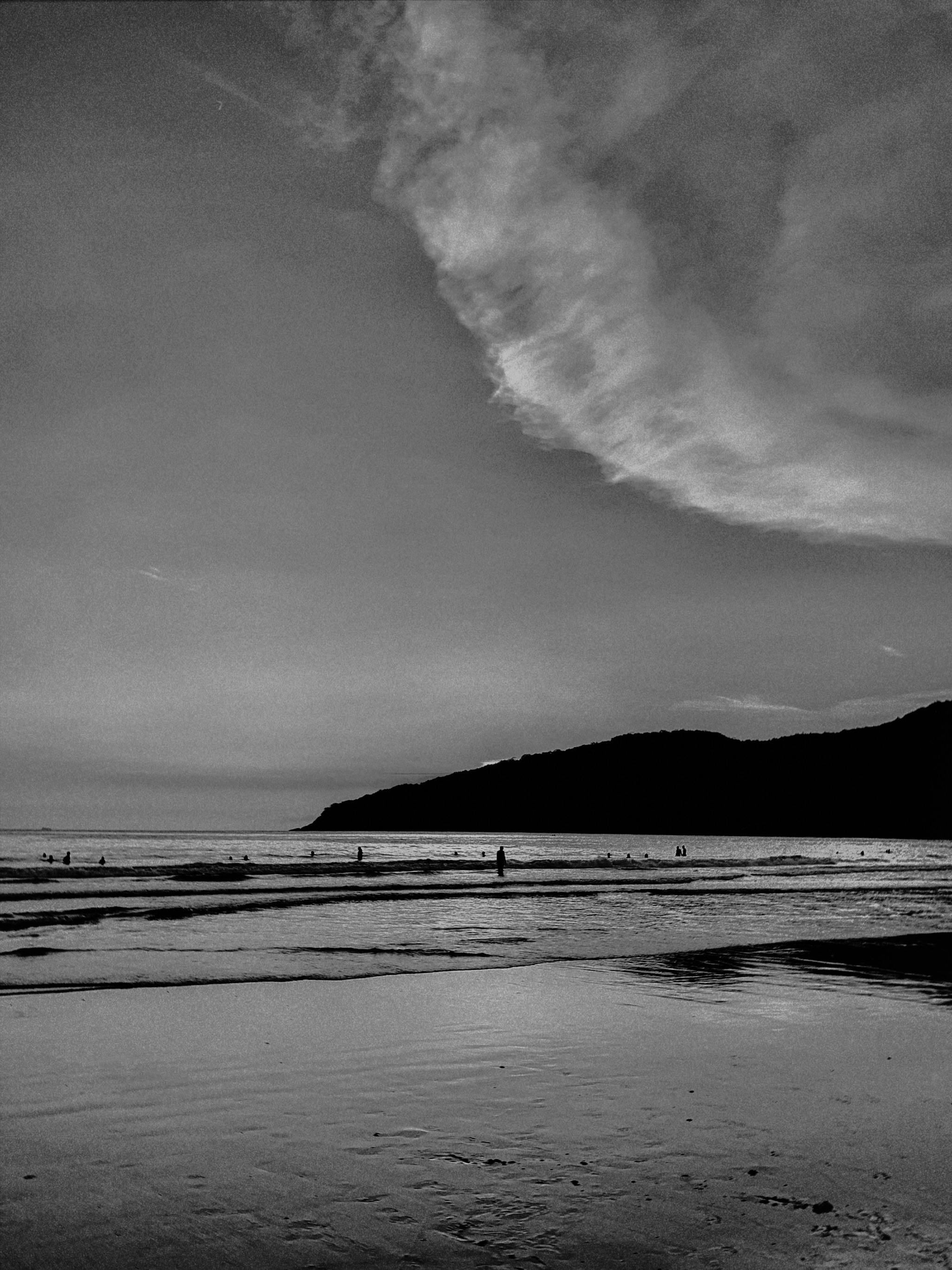 Black and white photo of a serene beach at sunrise with waves and distant silhouettes of people.