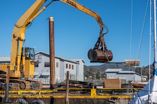 Yellow excavator with crane bucket at a construction site near buildings.