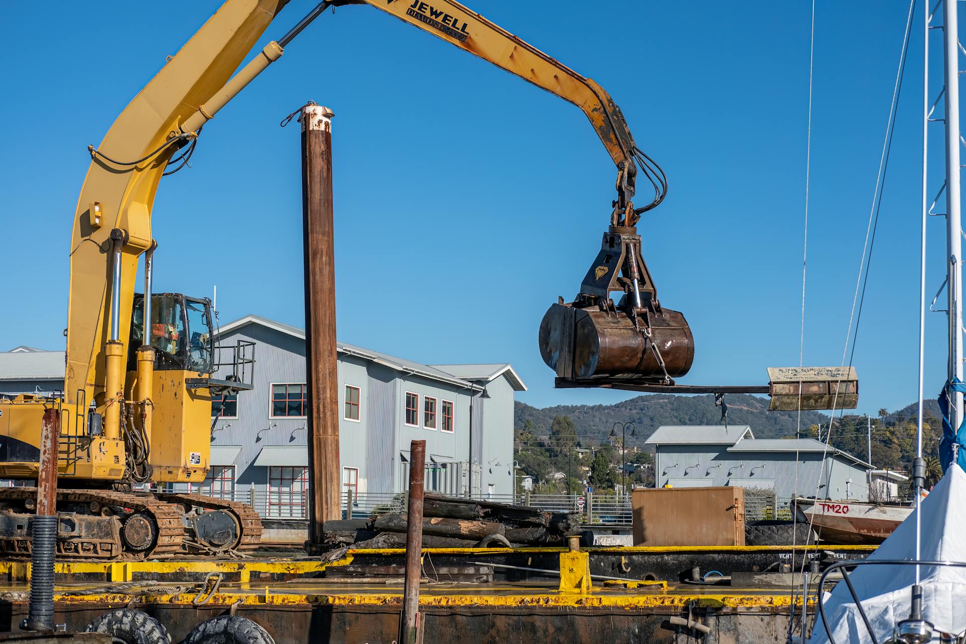 Yellow excavator with crane bucket at a construction site near buildings.