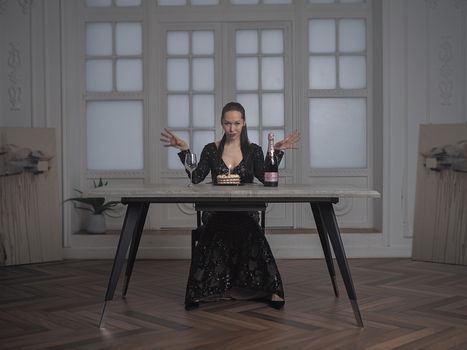 Woman in black dress sitting at table with cake and champagne, elegant setting.