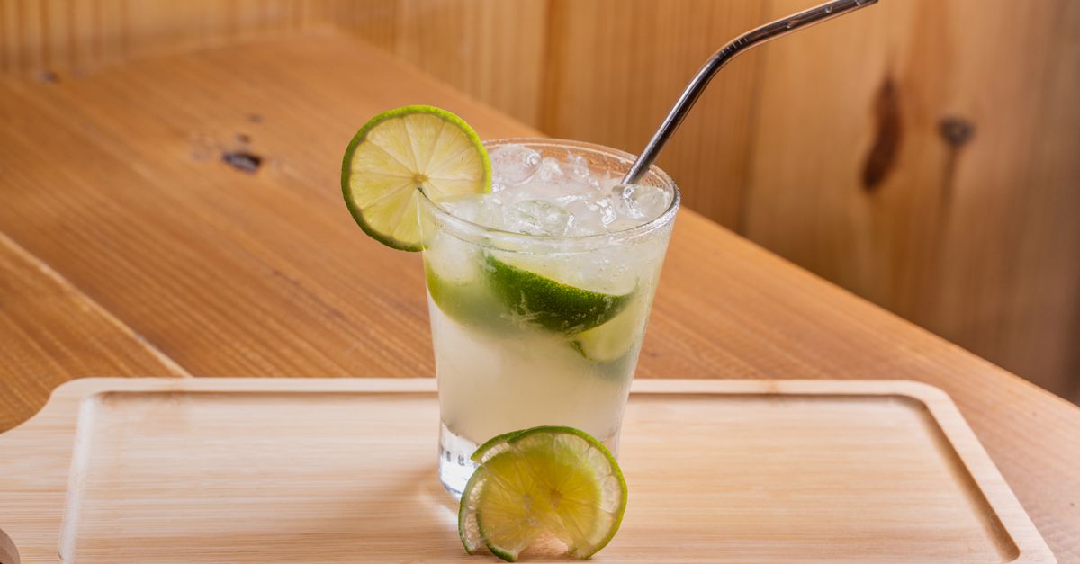 Glass of refreshing lime cocktail with ice and stainless steel straw on wooden tray.