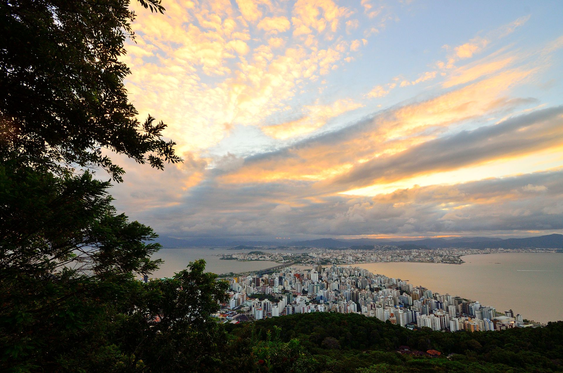 Stunning aerial view of Florianópolis skyline and ocean at sunset with vibrant clouds and lush greenery.