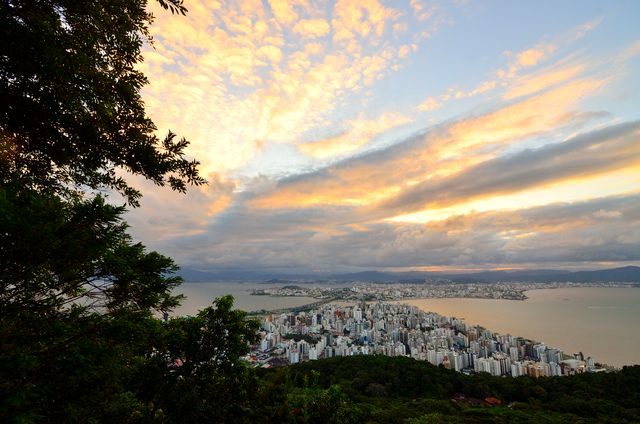 Stunning aerial view of Florianópolis skyline and ocean at sunset with vibrant clouds and lush greenery.