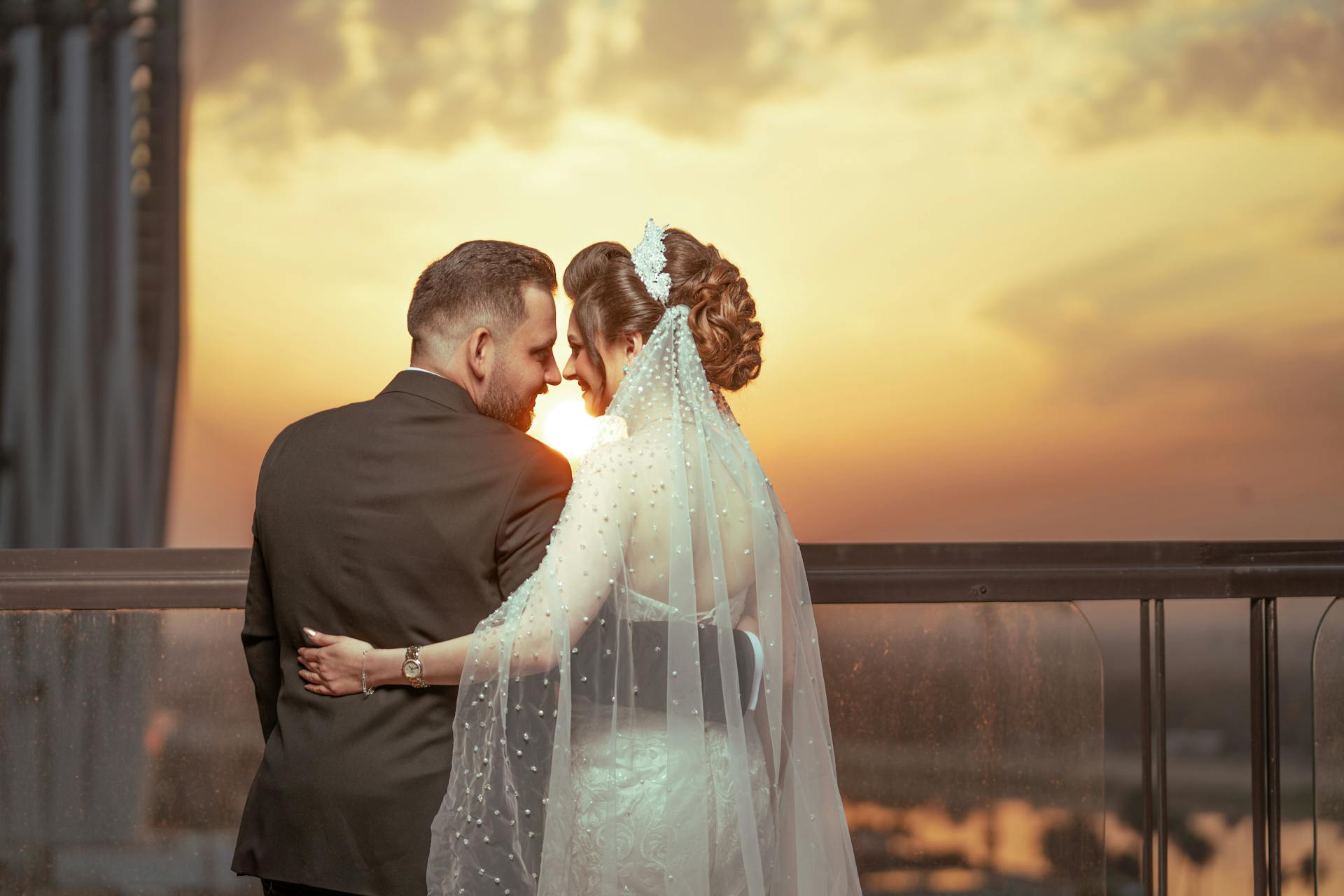 A couple embraces in wedding attire on a balcony with a stunning sunset backdrop.