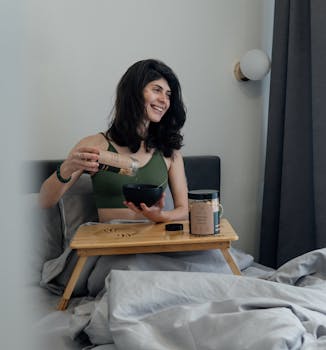 Caucasian woman in green top smiling, having breakfast in bed with a wooden tray.