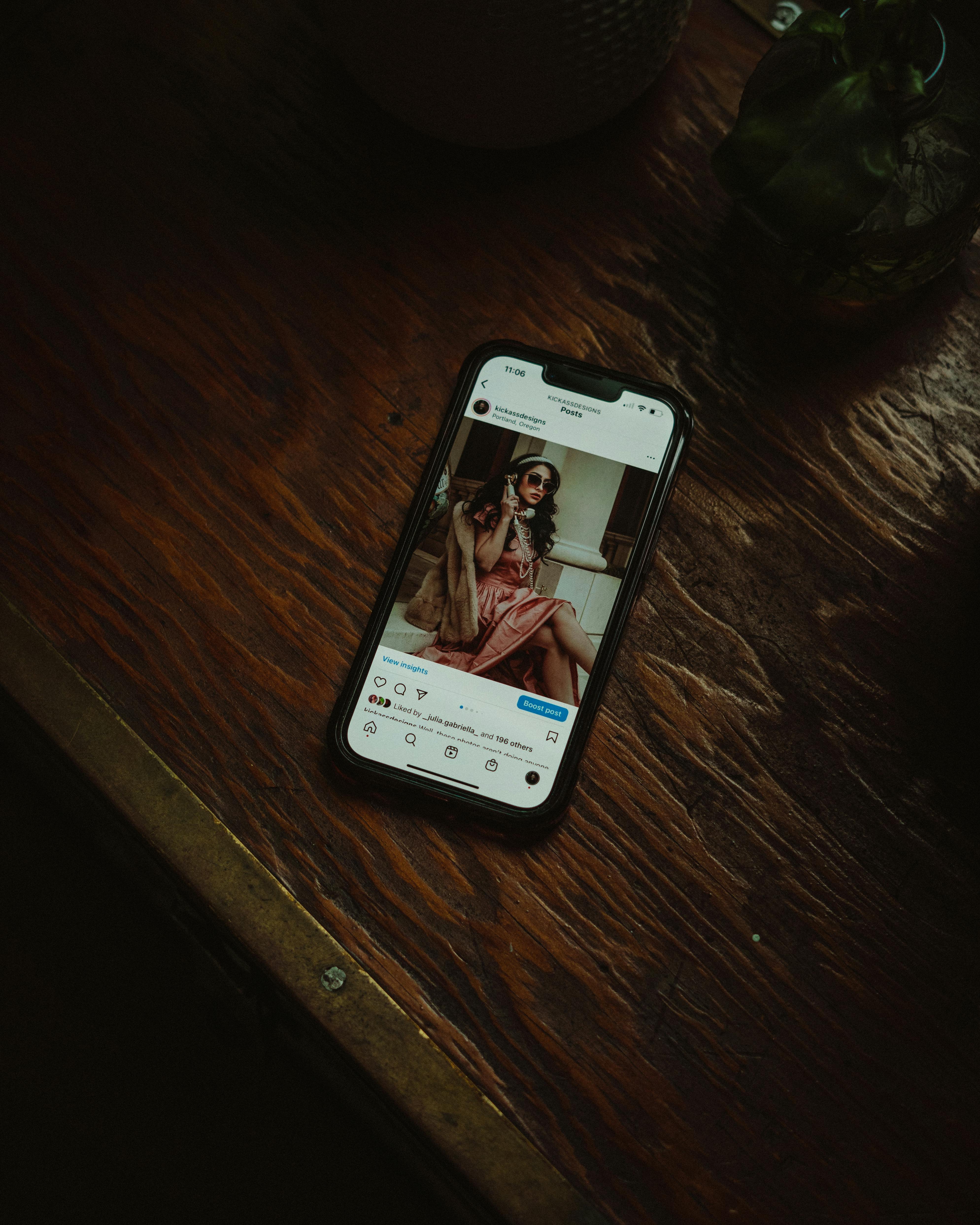 A smartphone on a wooden table shows a social media post with a stylish woman.