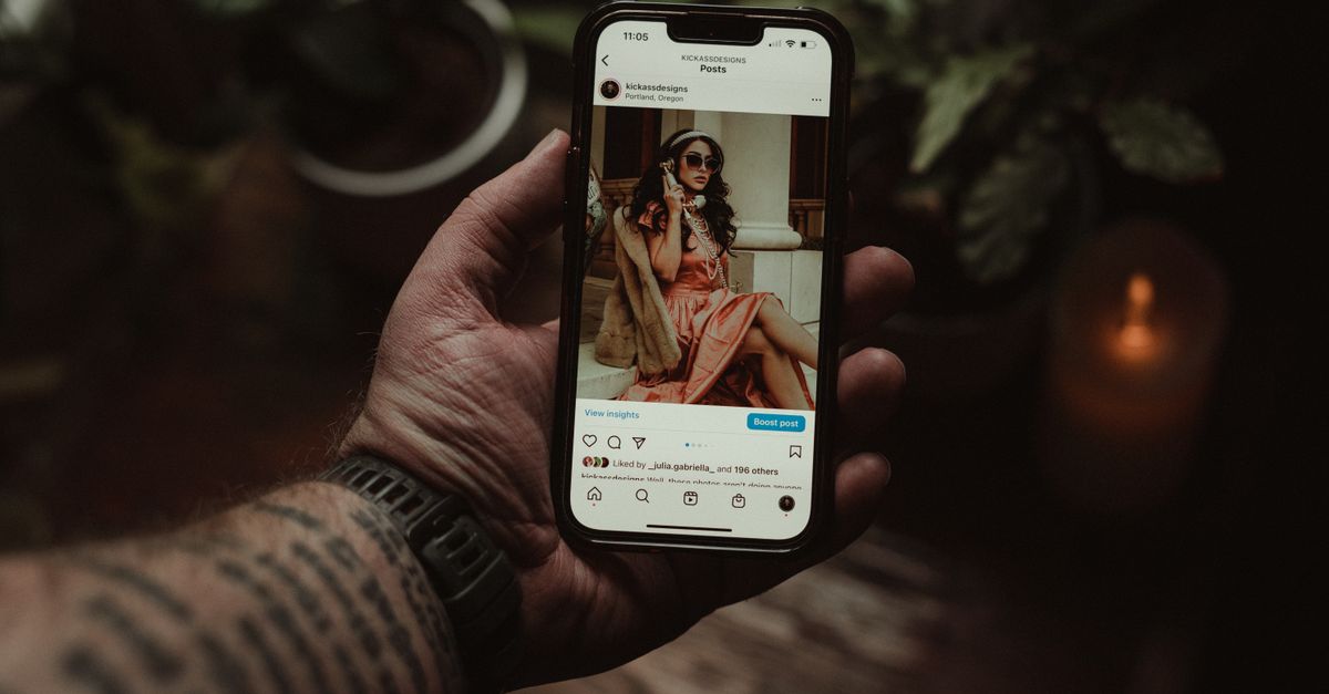 A tattooed hand holding a smartphone displaying an Instagram post indoors.