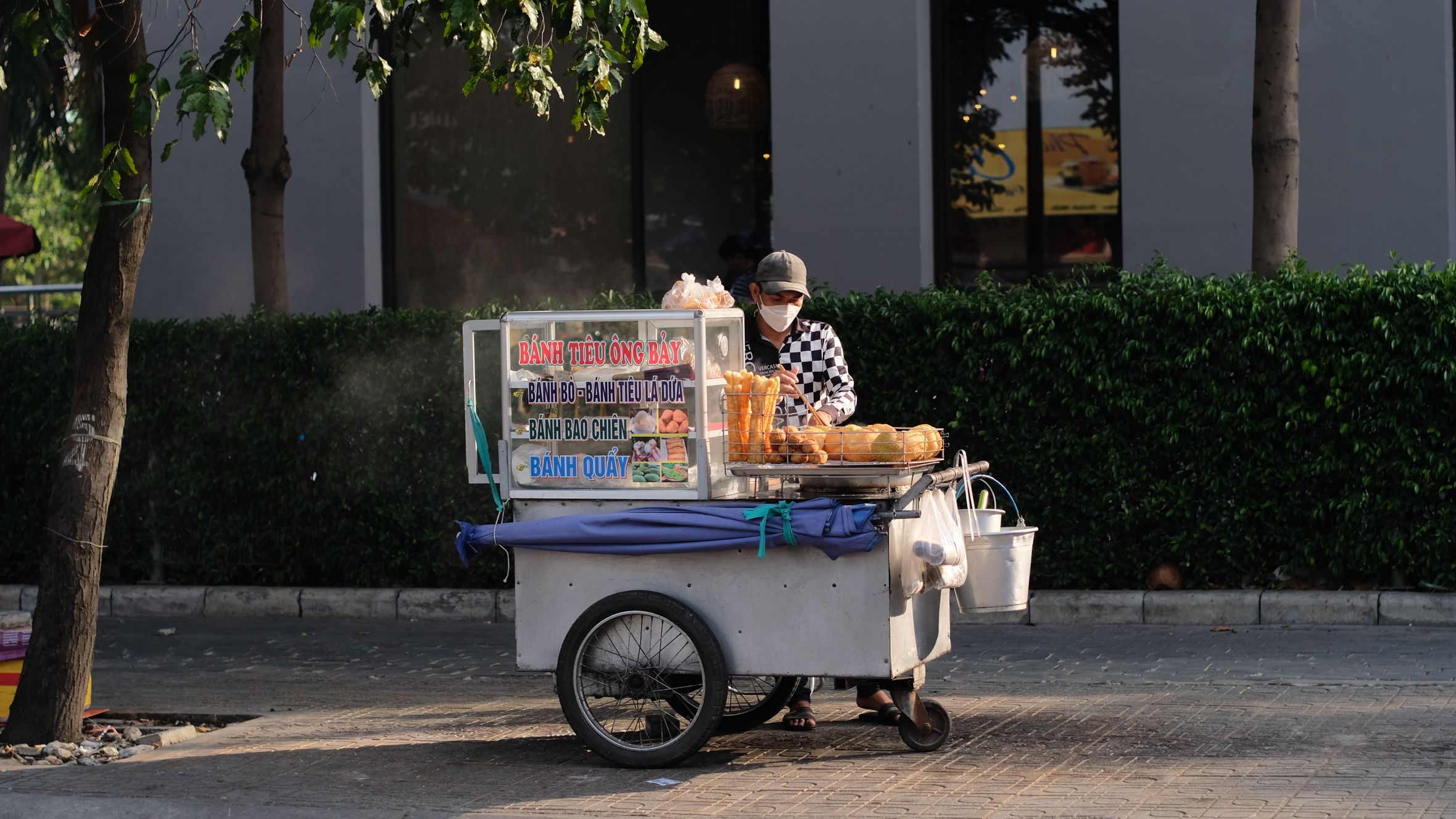 A street vendor with a cart selling bánh tiêu and other pastries on a sunny day.
