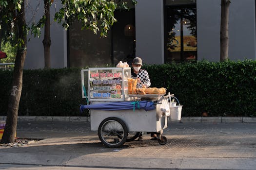 A street vendor with a cart selling bánh tiêu and other pastries on a sunny day.